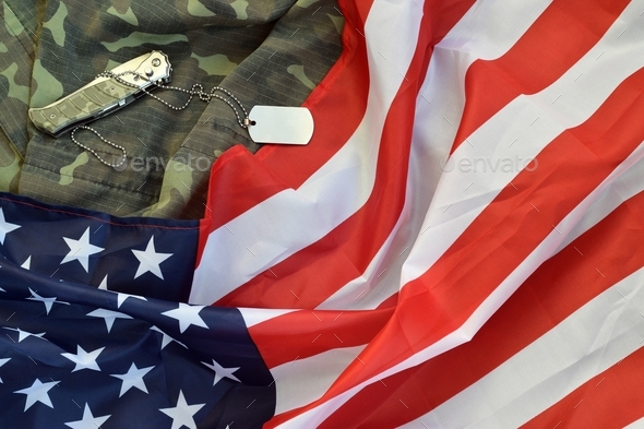 Army dog tag token and knife lies on Old Camouflage uniform and folded United States Flag