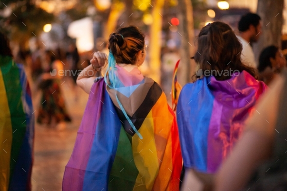 Young women from behind in rainbow flags at Pride party festival walking at night in the street