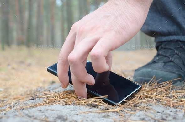 Male hand picking up a lost mobile phone from a ground in autumn fir wood path