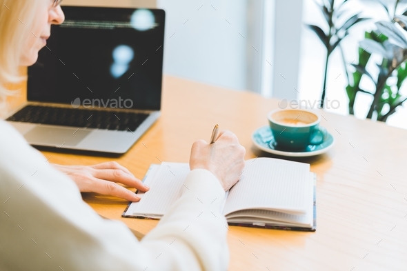Mature adult woman working online from cafe with coffee. Writing in notepad with laptop
