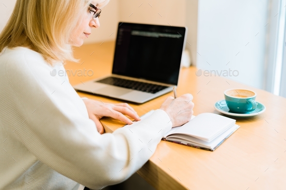 Mature adult senior woman working online from cafe with coffee. Writing in notepad with laptop