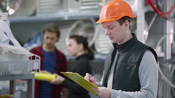 Confident Employee in Hard Hat Writing Register and Looking at Camera Smiling with Blurred Clients alt