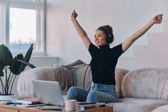 Excited young woman in headphones keeping arms raised while sitting near the laptop at home - Stock Photo - Images