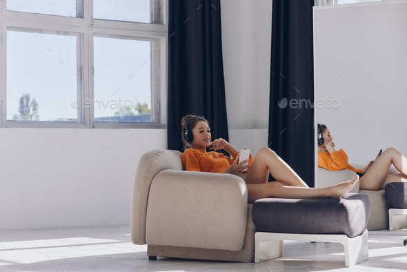 Attractive young woman in headphones using smart phone while relaxing in comfortable chair - Stock Photo - Images