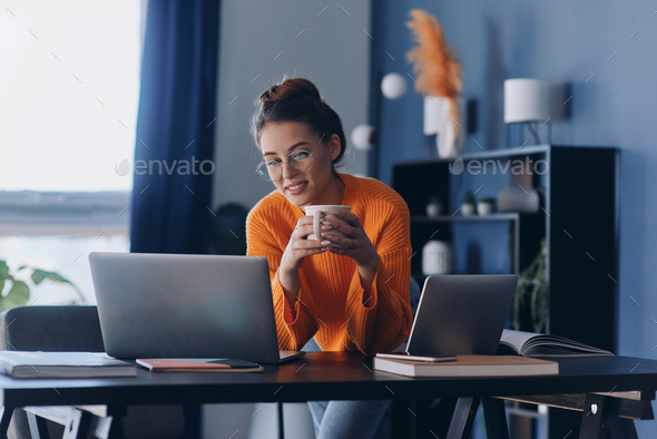Beautiful woman holding coffee cup and using laptop while sitting at her working place in office - Stock Photo - Images