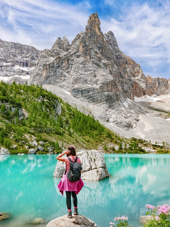 A young millennial woman in hiking clothes standing on the shore of an emerald lake under a mountain