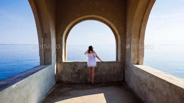 Woman standing under arched structure overlooking the sea. Minimalism, symmetry, travel, wanderlust.