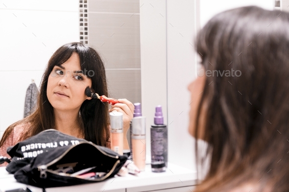 Young woman in front of bathroom mirror, doing makeup, using makeup brush.