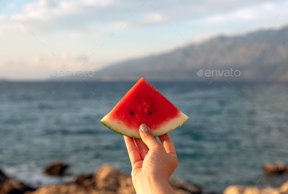 Personal perspective of woman holding a piece of watermelon on beach.