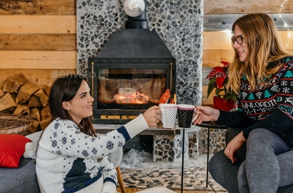 Two friends holding mugs with tea or mulled wine in front of fireplace in cozy cabin.