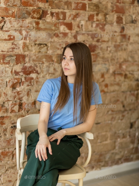 a girl in a blue t-shirt and green trousers sits on a chair in a room, studio, office