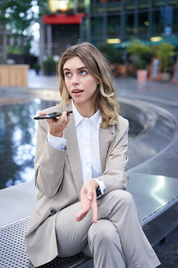 Smiling business woman record voice message, speaking into microphone on mobile phone, sitting near