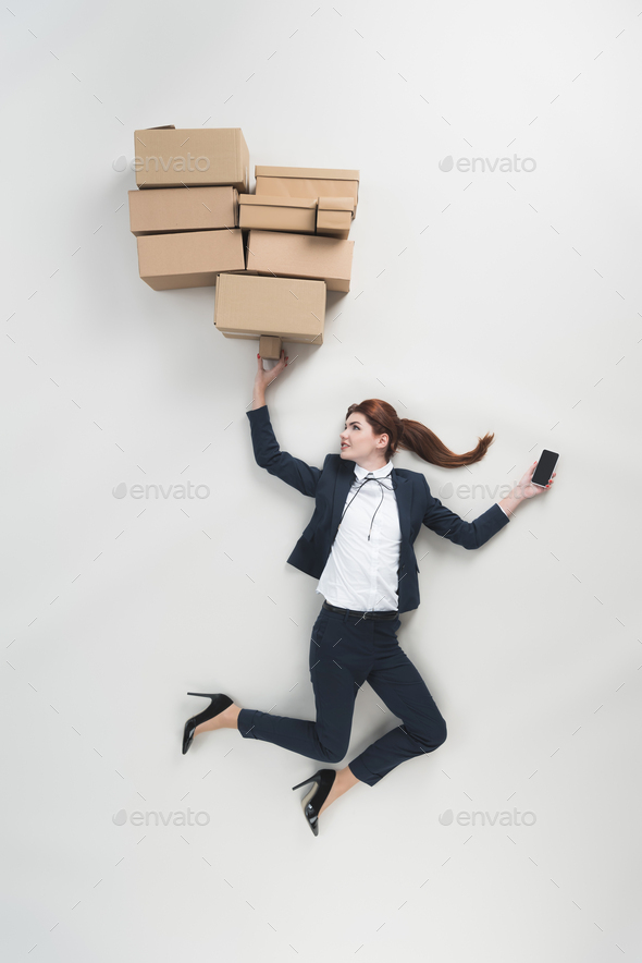 overhead view of businesswoman with smartphone holding pile of cardboard boxes isolated on grey