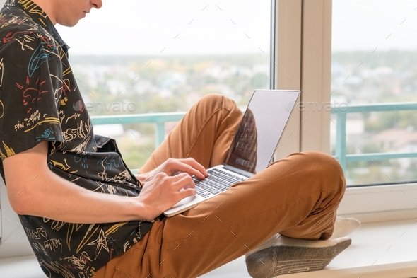 young man sits on the windowsill in the living room and uses laptop