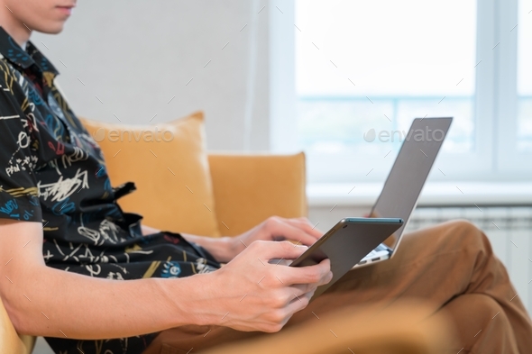 young man sitting on sofa in living room and using laptop and tablet