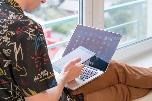 young man sits on the windowsill in the living room and uses laptop and tablet
