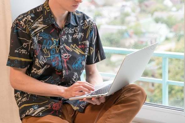 young man sits on the windowsill in the living room and uses laptop