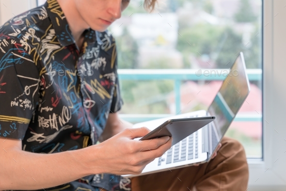 young man sits on the windowsill in the living room and uses laptop and tablet