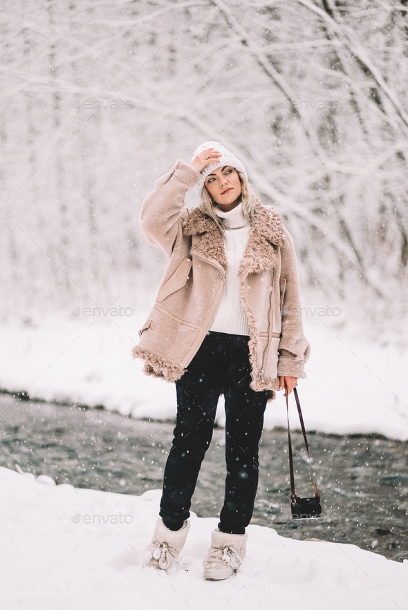 Young trendy woman in winter outfit posing under snowfall near