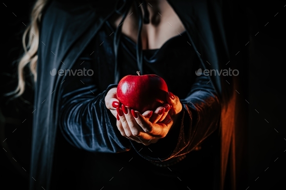 Witch woman in black offers red apple as symbol of temptation, poison. Fairy tale, white snow wizard