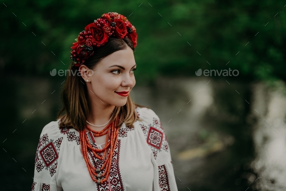 Pretty ukrainian woman in traditional embroidery vyshyvanka dress and red flowers wreath near river.
