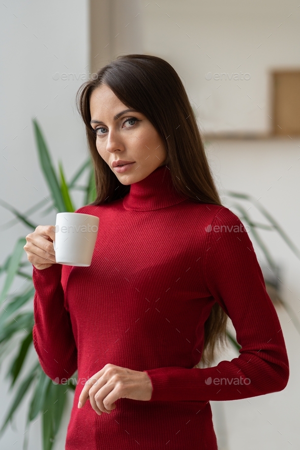 Pensive woman wear red turtleneck, looking at camera, holding white mug, drinking coffee or tea.