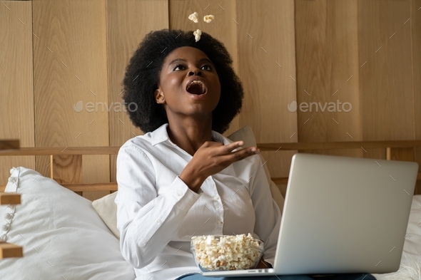 Happy Afro-American woman relaxing, toss popcorn up, watching movie. Lazy rest at home.