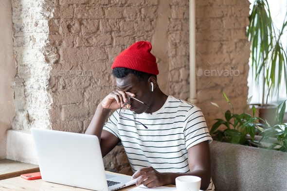 Afro-American man holding glasses, rubbing his eyes, feels tired after working on laptop in cafe