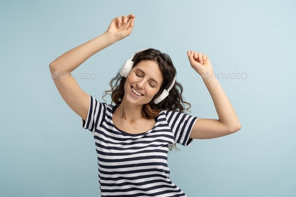 Woman in stripped t shirt wears wireless headphones, listens to music dancing with hands up isolated