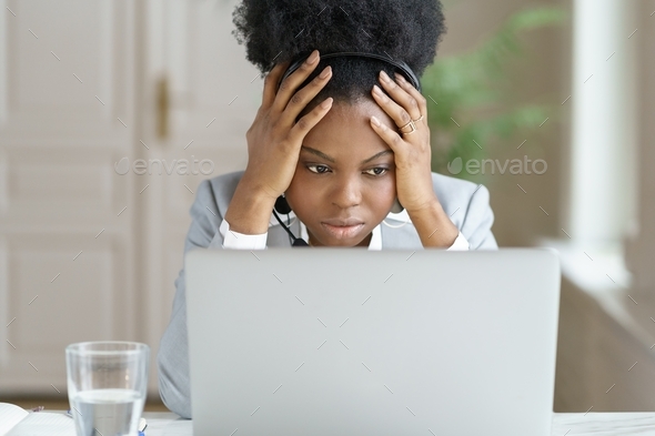 Tired annoyed afro woman wear headphones talking to a bad client looking at screen laptop in office