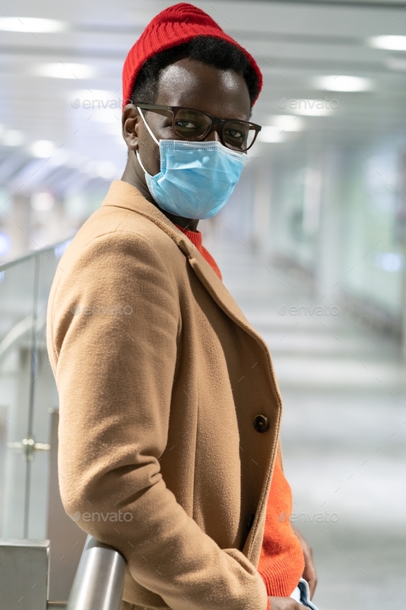 Trendy African hipster man looking at camera, standing in airport terminal, wear face mask. Covid-19