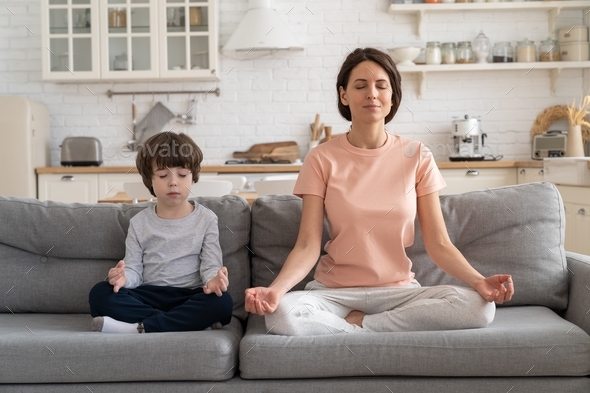 Mom with cute little son doing yoga exercise sit together on sofa at home teaching child to meditate