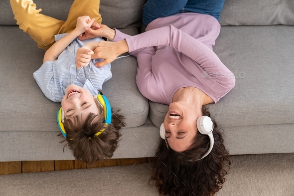 Happy mom and kid lying upside down on couch at home, laughing, tickle each other listening to music