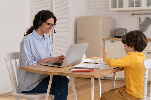 Mom work remotely with small son from kitchen. Young businesswoman home office with little boy draw