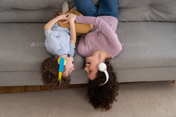 Happy mom and kid lying upside down on couch at home, laughing, tickle each other listening to music