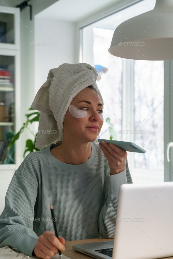 Woman with towel on head patches under eyes work from home on laptop records voice message on mobile