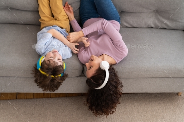 Smiling happy mother and kid lying on couch at home laughing tickling each other, listening to music