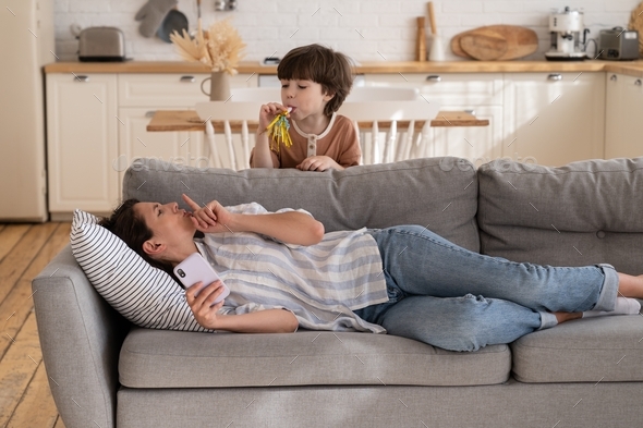 Stressed mom ask active son to calm. Tired mother try to rest after work on couch hold smartphone
