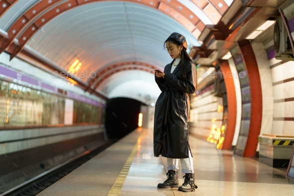 Trendy asian girl wait for train at subway platform check email or online schedule in smartphone