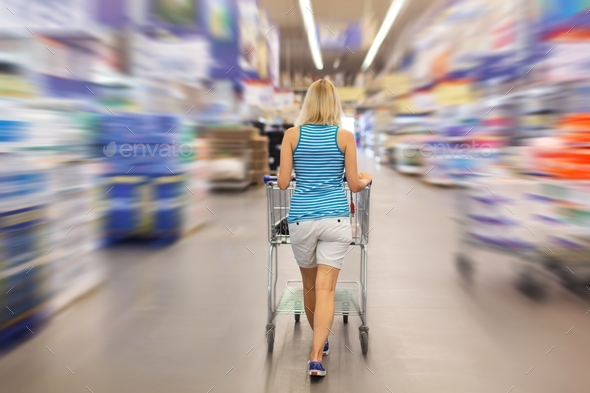 A young woman in a large supermarket pulls a cart between the aisles. Shopping in the hypermarket.