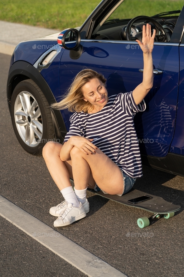 Excited middle age woman waving hand greeting friend sitting on longboard outside car in countryside