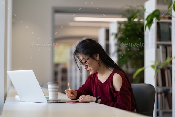 Nerdy Asian girl in glasses working on university research project in library or university