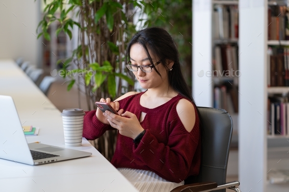 Student girl procrastinate in library with smartphone chat in social media not preparing for exam