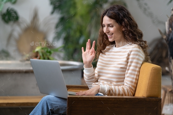 Italian woman waving hello, having video zoom call on laptop sitting in armchair in cozy home garden