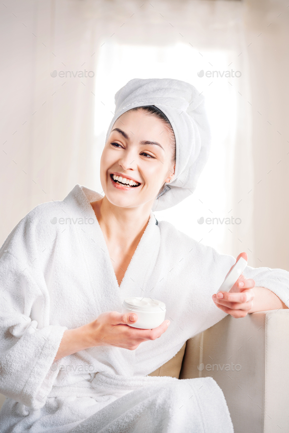 Laughing young woman in white bathrobe opening up a jar of face cream
