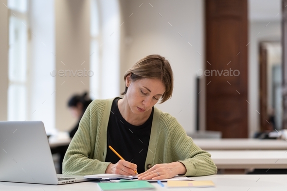 Middle-aged female student preparing for exam in public library while sitting at table with laptop