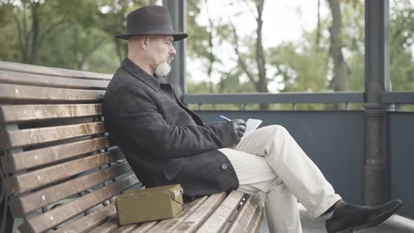 Side View of Elegant Undercover Agent or Spy Sitting on Bench in Cloudy Spring or Autumn Park and alt
