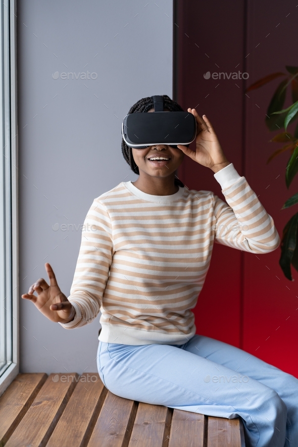 Joyful afro-american woman in VR helmet enjoying shopping in virtual reality while sitting indoors