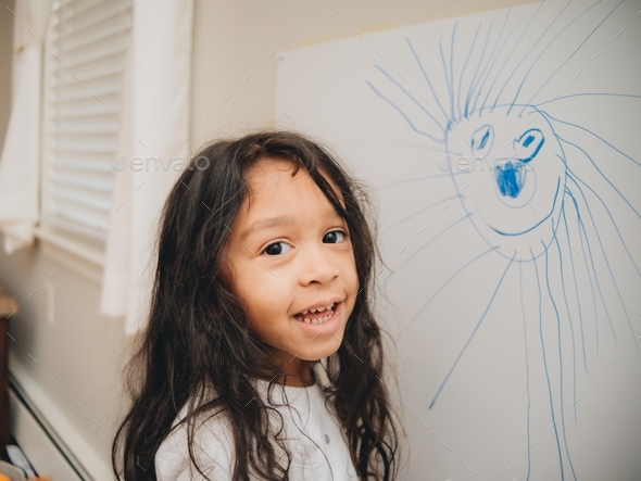 Diverse pre school girl at home drawing funny characters and faces on dry erase board
