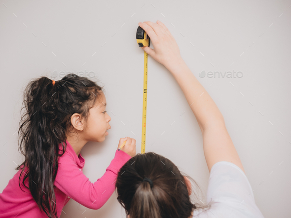 Mother and diverse daughter at home using tape measure to measure height and write it on wall